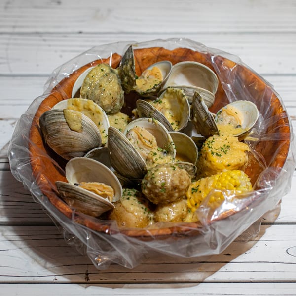 Steamed clams, potatoes, and corn on the cob in a wooden bowl, covered in a creamy sauce.