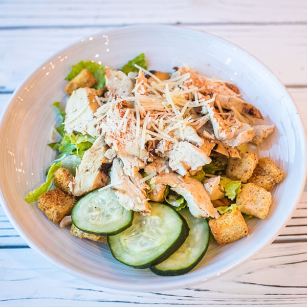 Chicken Caesar salad with shredded cheese, croutons, and cucumber slices in a white bowl.