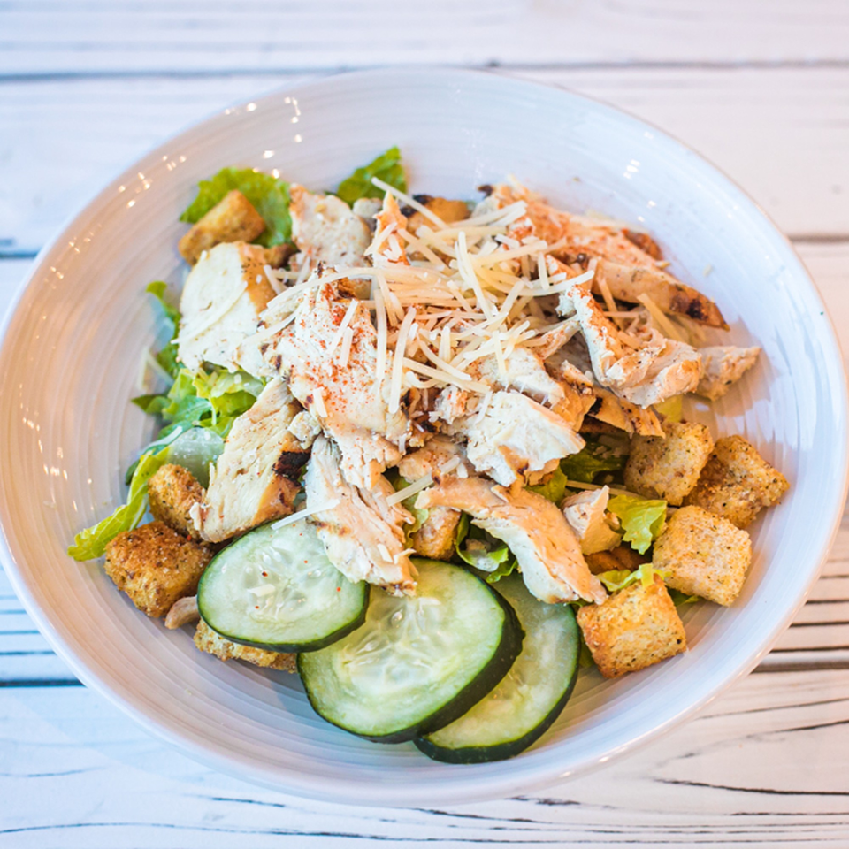 Chicken Caesar salad with shredded cheese, croutons, and cucumber slices in a white bowl.