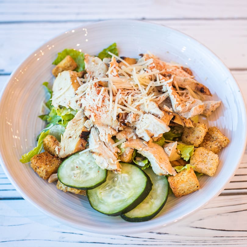 Chicken Caesar salad with shredded cheese, croutons, and cucumber slices in a white bowl.