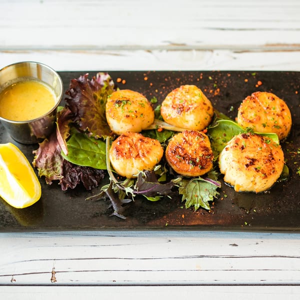 Six seared scallops on a bed of greens with a side of lemon and butter sauce.