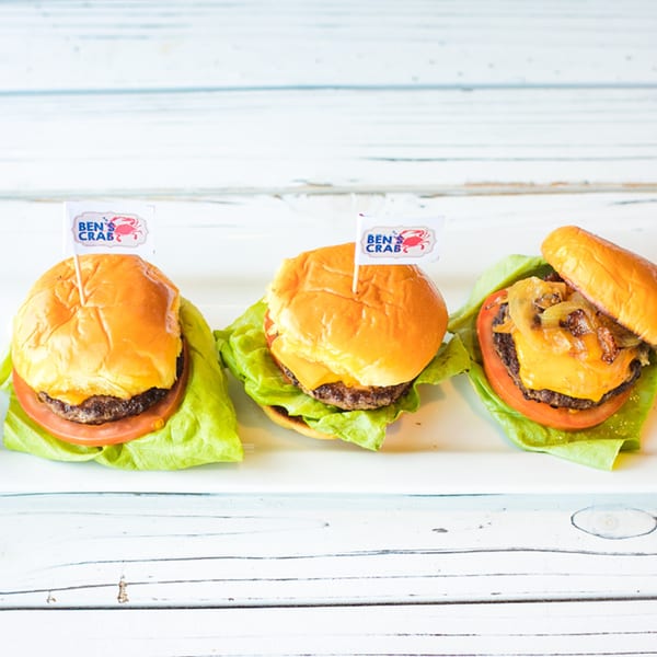 Three Ben's Crab mini cheeseburgers with lettuce, tomato, and one with grilled onions on a white surface.