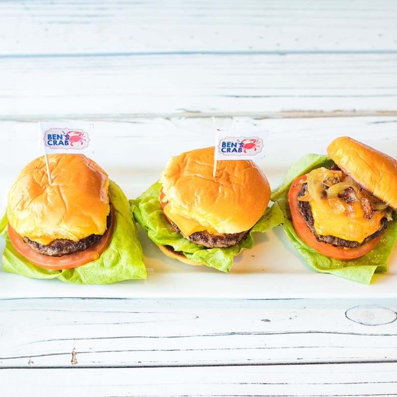 Three Ben's Crab mini cheeseburgers with lettuce, tomato, and one with grilled onions on a white surface.