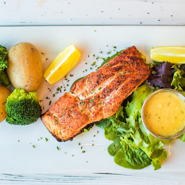 Grilled salmon with potatoes, broccoli, lemon wedges, and a side of sauce on a white plate.