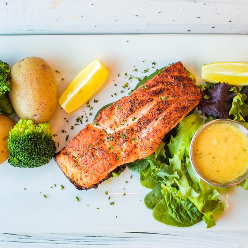 Grilled salmon with potatoes, broccoli, lemon wedges, and a side of sauce on a white plate.