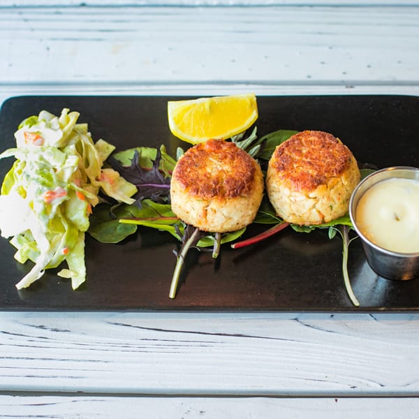 Two Ben's Crab crab cakes with coleslaw, a lemon wedge, and dipping sauce on a black plate.