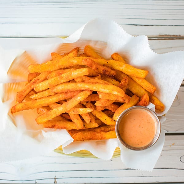 A basket of golden-brown seasoned fries with a side of creamy orange dipping sauce on a white wooden table.